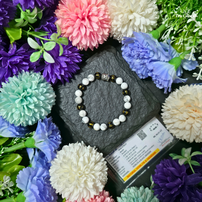 A beaded bracelet with white marbled beads, black beads with gold accents, and a small silver-toned Buddha bead, displayed on a dark slate stone surrounded by colorful artificial flowers.