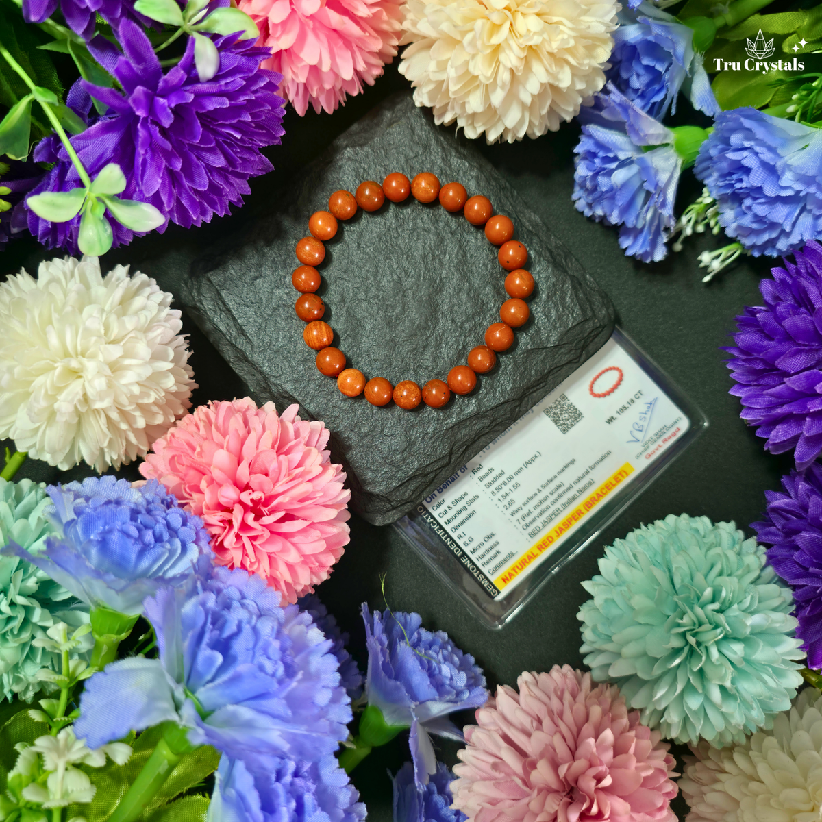 A circular beaded bracelet made of Red jasper gemstone beads sits on a dark slate tile next to a clear display card. The scene is styled with colorful artificial flowers in pink, purple, blue, white, and mint around the bracelet.