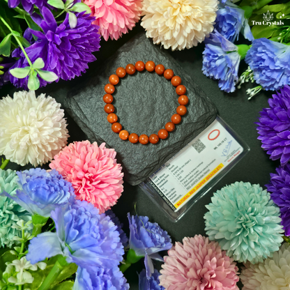 A circular beaded bracelet made of Red jasper gemstone beads sits on a dark slate tile next to a clear display card. The scene is styled with colorful artificial flowers in pink, purple, blue, white, and mint around the bracelet.