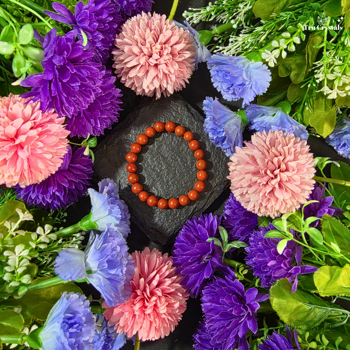A circular beaded bracelet made of Red jasper gemstone beads sits on a dark slate tile next to a clear display card. The scene is styled with colorful artificial flowers in pink, purple, blue, white, and mint around the bracelet.