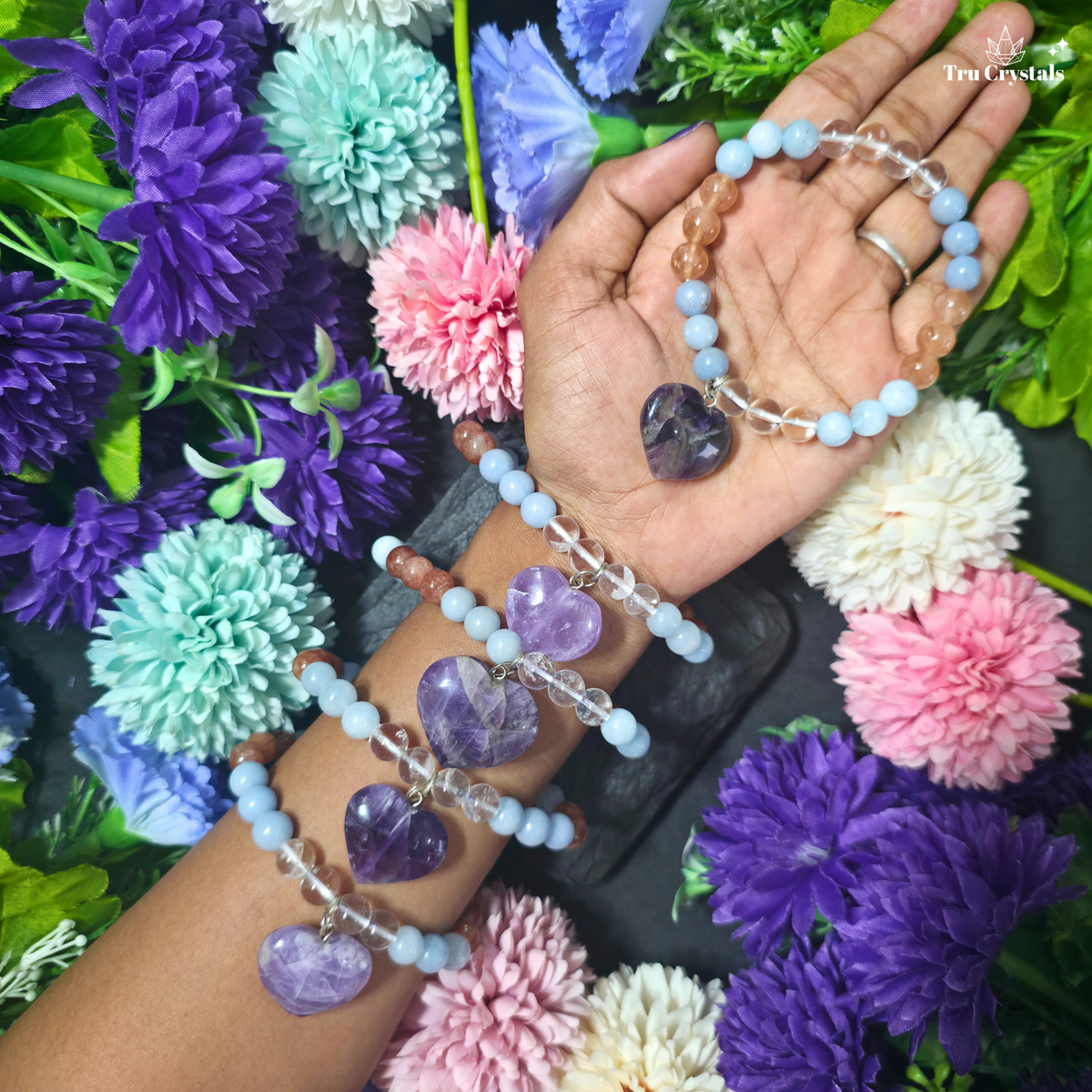 Three beaded crystal bracelets with heart-shaped amethyst pendants are displayed on a person's wrist and held in their hand, surrounded by colorful artificial flowers. The bracelets feature light blue, peach, and clear beads.