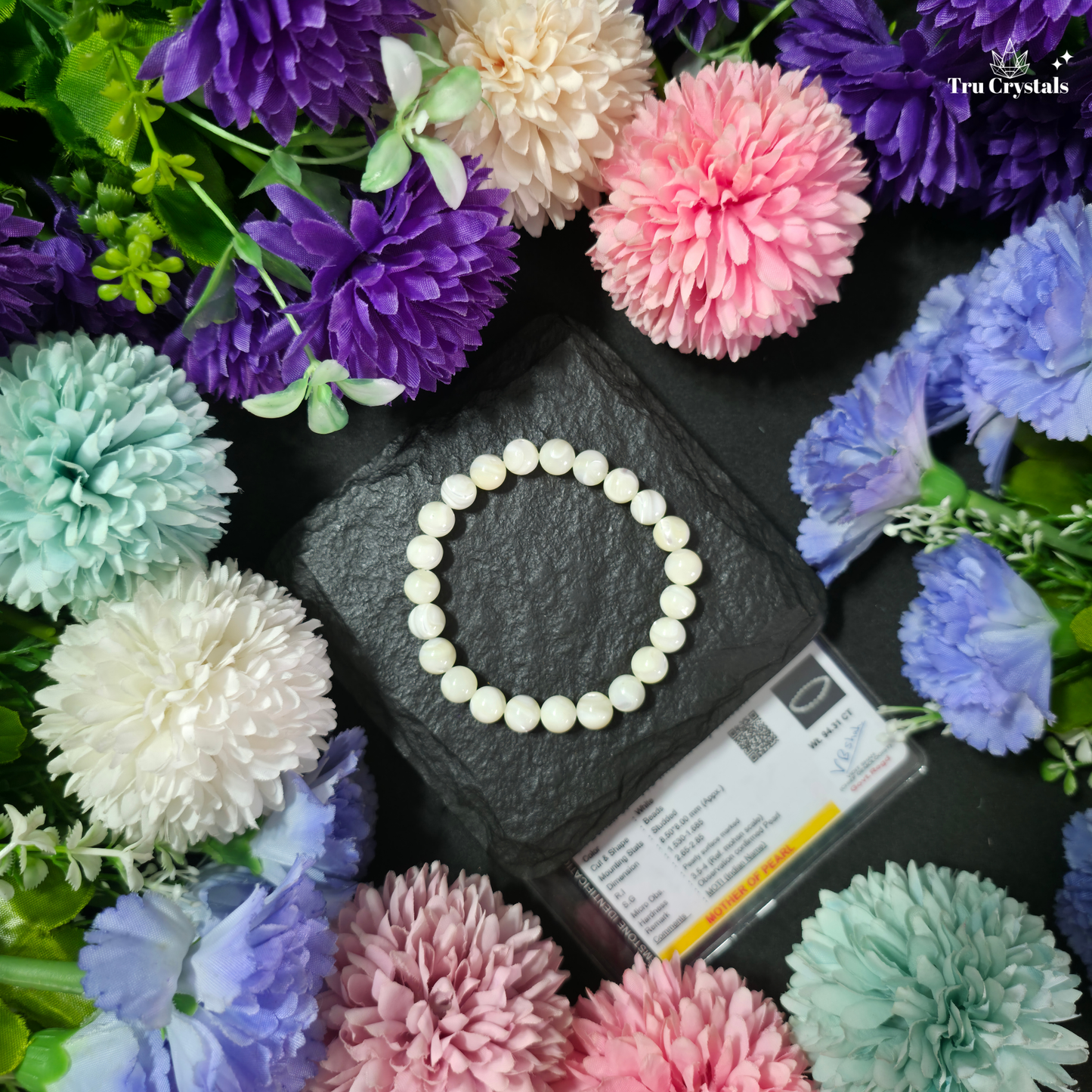 A beaded bracelet made of round mother-of-pearl is displayed on a dark slate block, surrounded by colorful artificial flowers in pink, purple, blue, and mint. A small product card is visible at the bottom right, and the Tru Crystals logo appears in the top right corner.