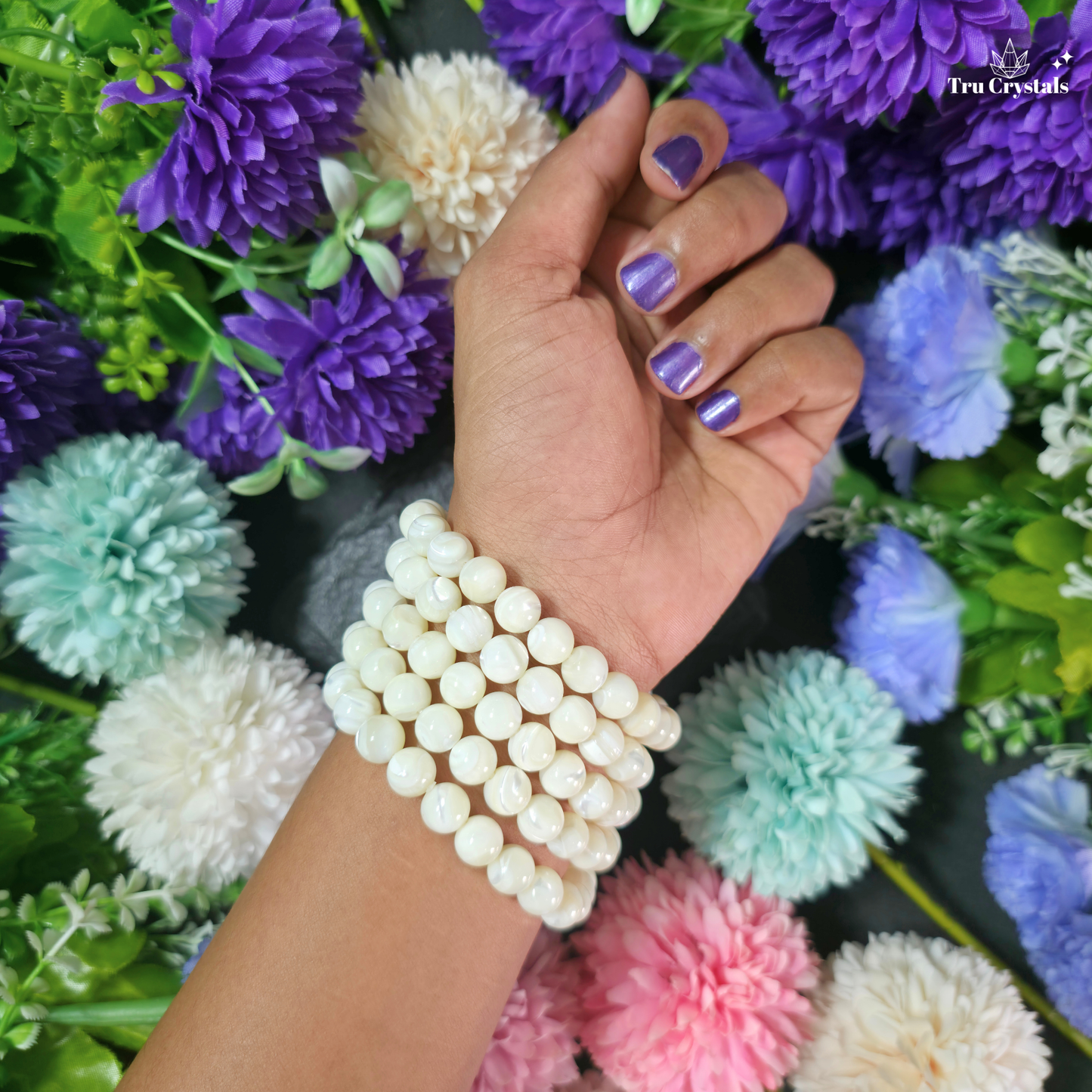 A close-up of a person's hand and wrist wearing a stack of cream-colored faux pearl beaded bracelets, set against a background of colorful artificial flowers.