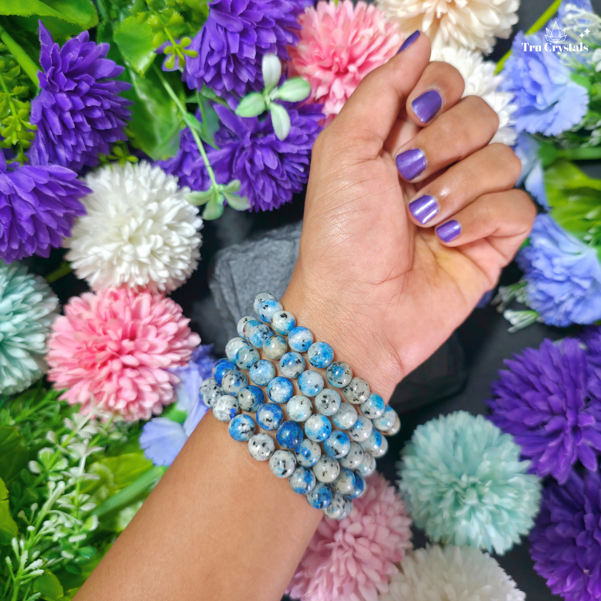A close-up of a person's hand wearing a stack of three beaded bracelets made of blue and white marbled stone beads, set against a background of colorful artificial flowers.