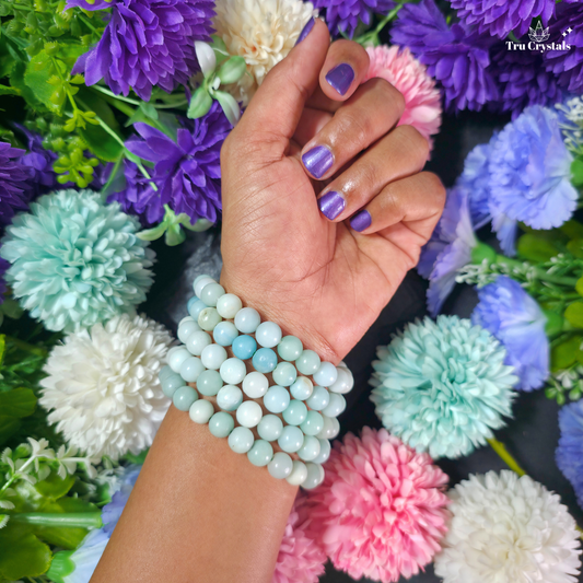 A close-up of a person's hand and wrist wearing a stack of light blue and white beaded  amazonite bracelets, surrounded by colorful faux flowers. The brand name 'Tru Crystal' is visible in the top right corner.