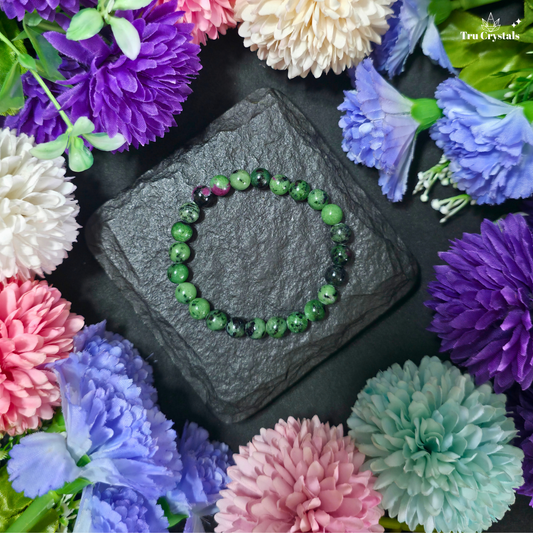 A beaded stretch bracelet made of Ruby zoistite beads, displayed on a dark slate stone surrounded by colorful artificial flowers. The brand name 'True Crystals' is visible in the top right corner.