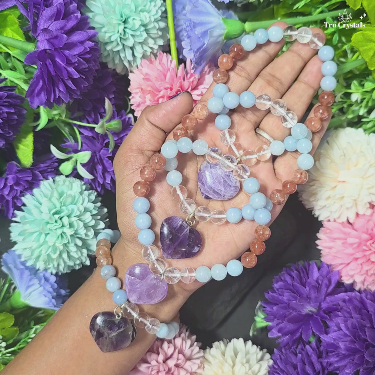 A multicolored stone bracelet presented on a white platform with a purple background featuring the logo 'Tru Crystals'. The bracelet composed of Aquamarine, sunstone, clear quartz,angelite and amethyst round beads and one large amethyst crystal at the center.