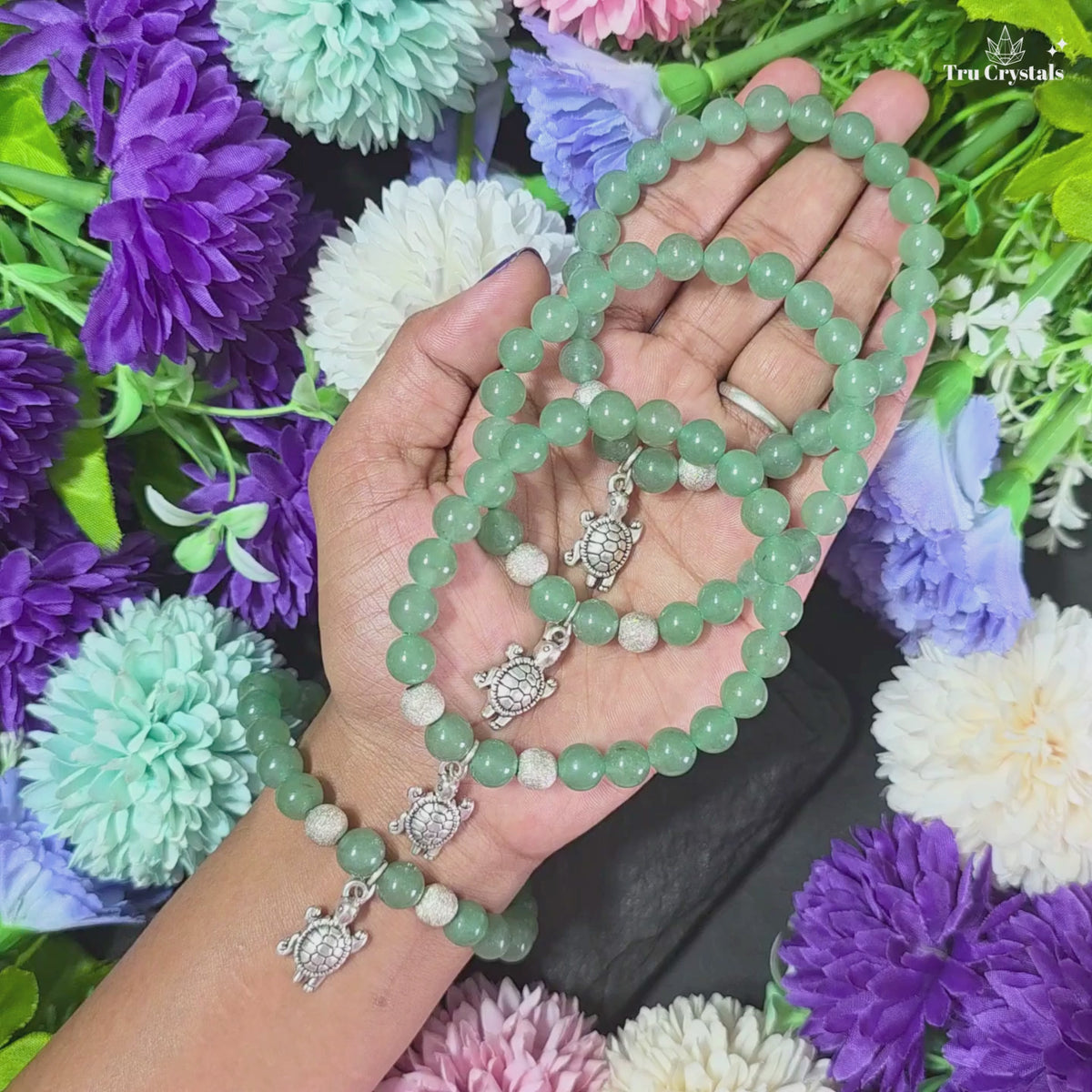 A Green Aventurine crystal bracelet with a tortoise charm placed on a dark stone, surrounded by colorful flowers.