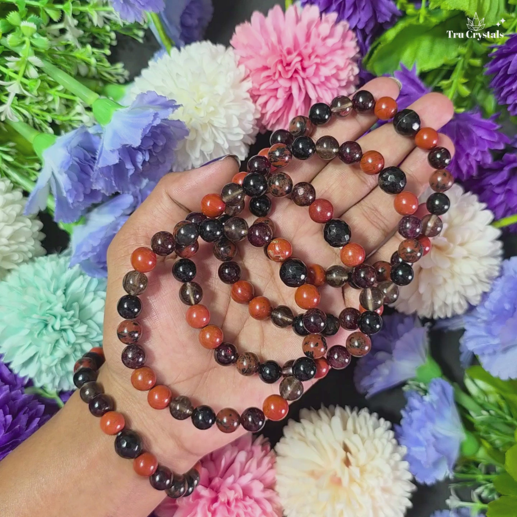 A bracelet made with beads of Red Jasper, Garnet, Smoky Quartz, Black Tourmaline, and Mahogany Obsidian, displayed on a dark stone surface surrounded by colorful flowers.