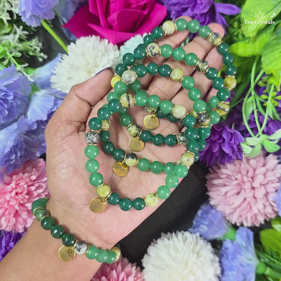 A green numerology bracelet with green jade, green aventurine, and serpentine stones, featuring a 'Be TRU' engraving on a black stone charm, displayed on a dark background with purple flowers.