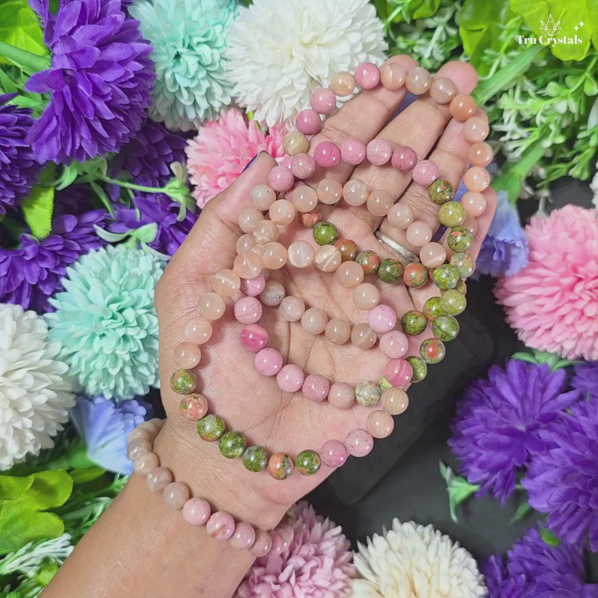 A multicolored gemstone bracelet containing moonstone, rhodochrosite and unakite displayed on a wooden surface with flowers and a colorful container in the background.
