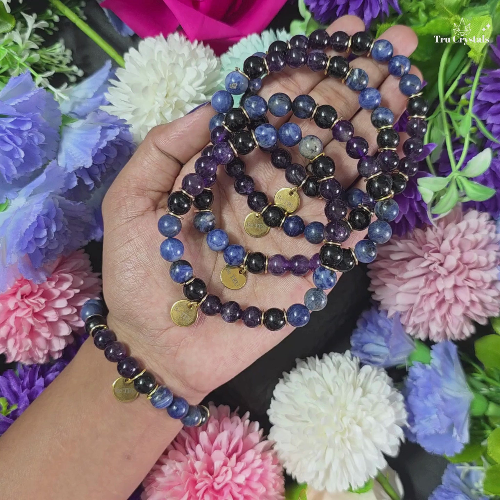 A bracelet with Sodalite, Amethyst, and Black Tourmaline  beads, featuring a 'Be TRU' engraved charm, displayed on a textured background with flowers.
