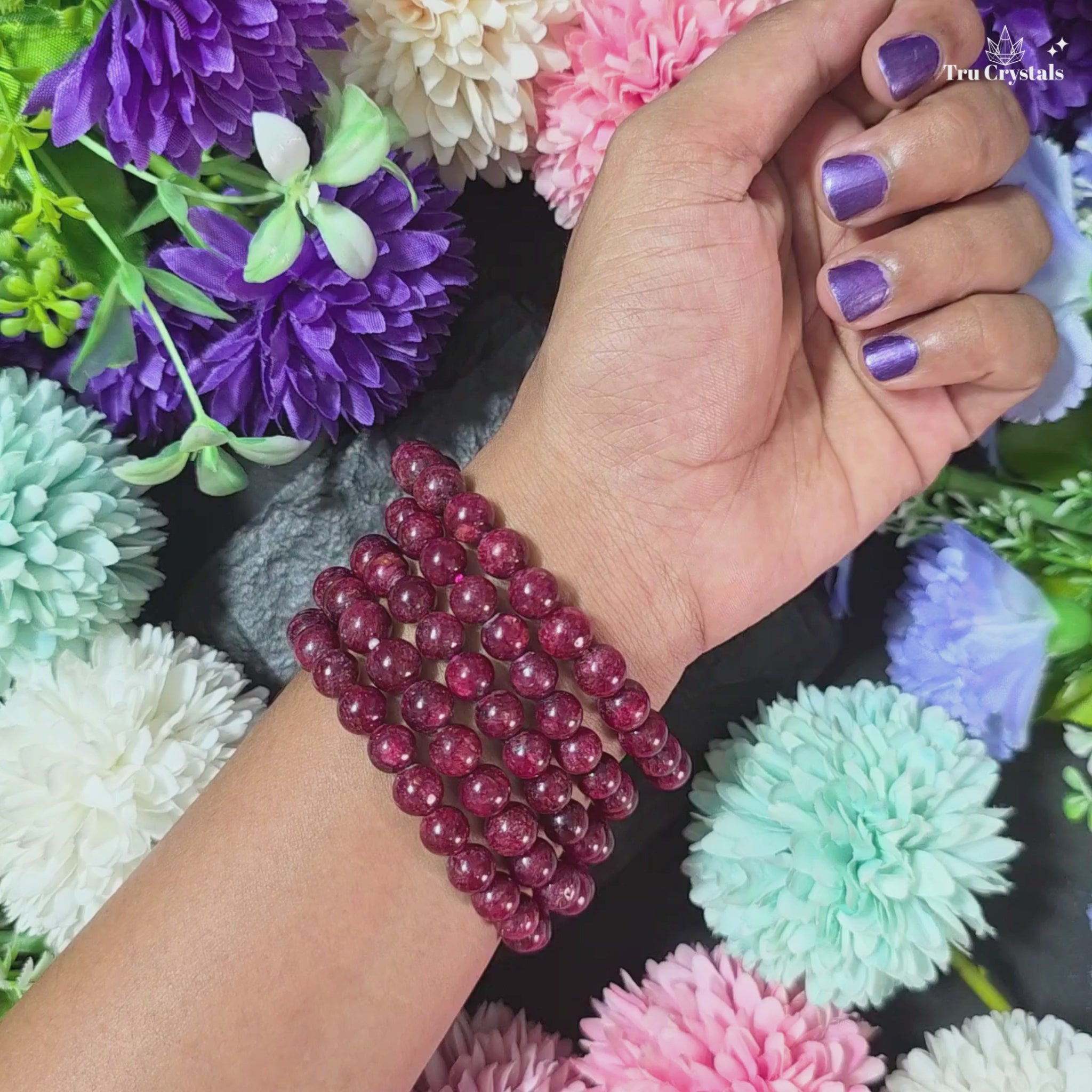 Natural Red  Ruby beaded bracelet on a dark stone surface surrounded by purple and blue flowers.