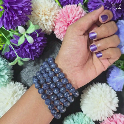 A beaded bracelet made with natural blue sapphire gemstones, displayed on a gray stone with purple and blue flowers around it