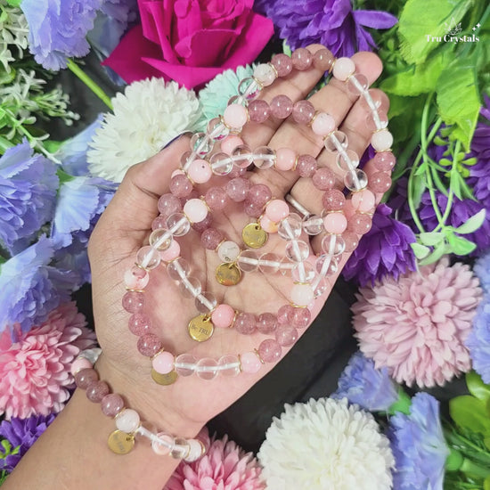 A  numerology bracelet for 6 and venus planet with Clear quartz , strawberry quartz and pink opal stones, featuring a 'Be TRU' engraving on a black stone charm, displayed on a dark background with purple flowers.