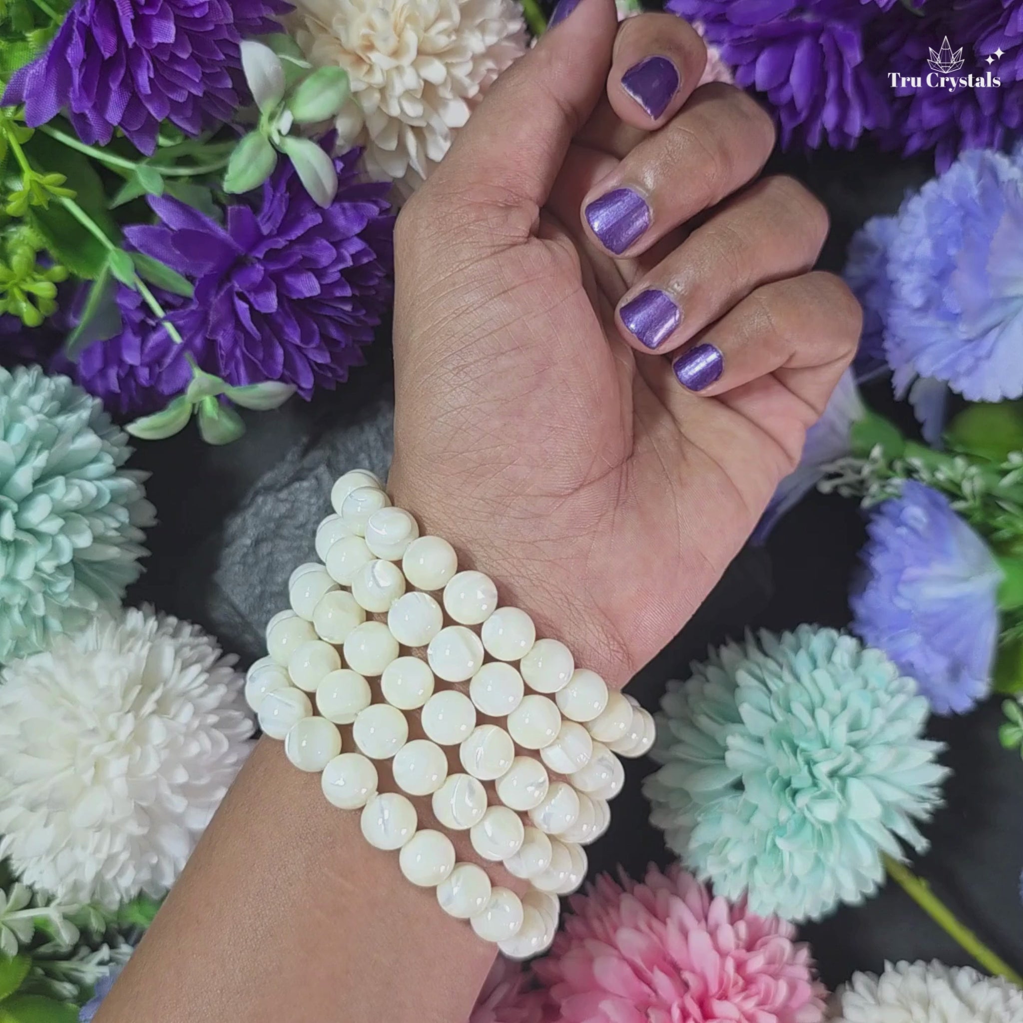A bracelet made of mother-of-pearl beads  for Calming Energy & Inner Wisdom displayed on a dark surface surrounded by colorful flowers.
