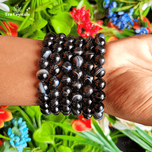 A black Sulemani Hakik Agate bracelet displayed on a grey surface surrounded by colorful flowers, with a certification card in the background.